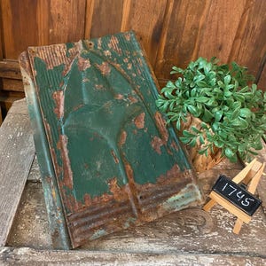 May include: Weathered, green metal architectural salvage with a stylized design. The metal shows signs of rust and wear, adding to its vintage appeal. A small chalkboard with the number "1745" is displayed nearby. A small green plant is also in the image.