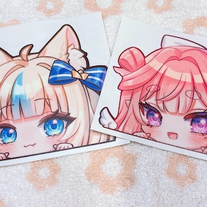May include: Two anime-style character stickers. One features a blonde character with blue accents and a blue bow. The other has pink hair and a pink bow. Both have large eyes and are peeking over a white surface.