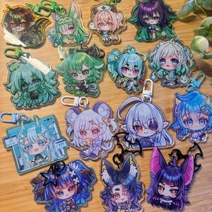 Peut inclure: Une collection de quinze porte-clés en acrylique représentant divers personnages de style chibi avec des cheveux aux couleurs vives, notamment des nuances de vert, de bleu et de violet, chacun attaché à un fermoir en plastique coloré en forme d'étoile ou de chauve-souris.