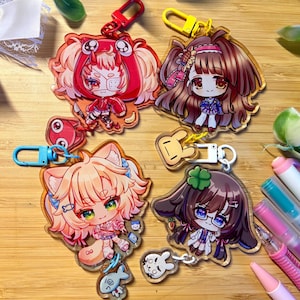May include: Four acrylic keychains featuring chibi-style anime characters with colorful hair and outfits. Each keychain includes a secondary smaller charm, such as a fish, a rabbit, or a creature, attached by a plastic clasp in red, yellow, blue, or white.