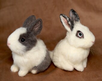 May include: Two small, white and gray, needle felted rabbits. One rabbit is sitting with its head turned to the right, while the other rabbit is sitting with its head turned to the left. Both rabbits have black eyes and pink noses.