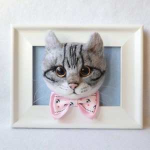 May include: A gray and white felted cat head with black eyes and a pink bow tie with white anchors. The cat head is framed in a white frame.