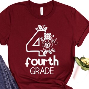 K&ouml;nnte beinhalten: Burgunderfarbenes T-Shirt mit wei&szlig;er Grafik. Die Grafik zeigt die Zahl 4, florale Akzente und die Worte "fourth GRADE" in fetter Schrift. Das T-Shirt ist gefaltet, eine Jeans ist in der Ecke sichtbar.