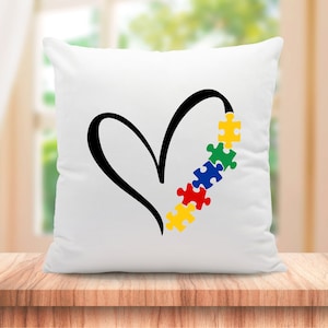 Puede incluir: Almohada blanca con un dise&ntilde;o de coraz&oacute;n negro y una fila de piezas de rompecabezas de colores que forman el lado derecho del coraz&oacute;n. Las piezas del rompecabezas son amarillas, verdes, azules, rojas y amarillas.