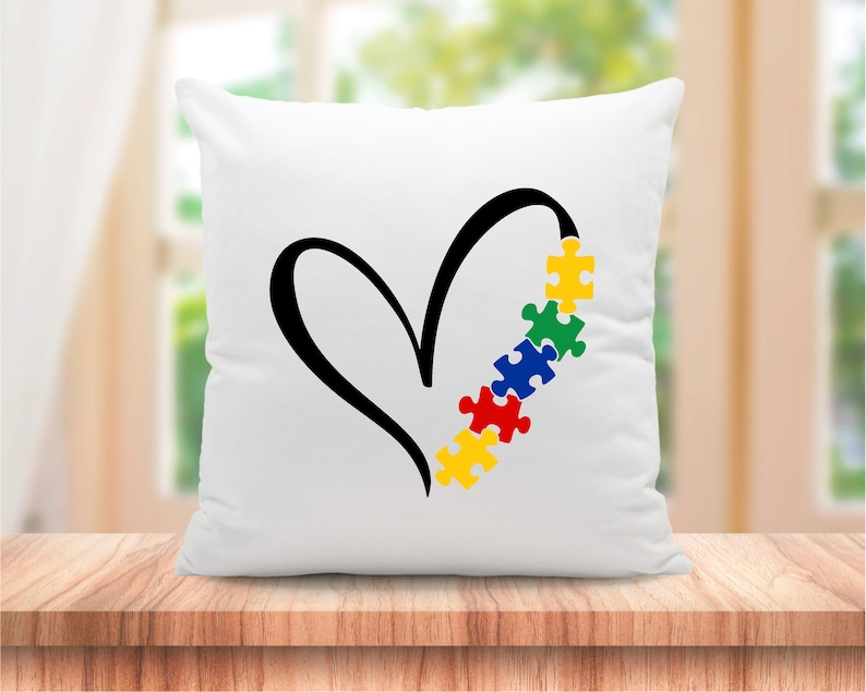 Puede incluir: Almohada blanca con un dise&ntilde;o de coraz&oacute;n negro y una fila de piezas de rompecabezas de colores que forman el lado derecho del coraz&oacute;n. Las piezas del rompecabezas son amarillas, verdes, azules, rojas y amarillas.