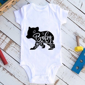 May include: White baby onesie with a black bear silhouette design. The bear is filled with floral patterns and the word "Baby" in a cursive font. The onesie is made of soft material and has snap closures.