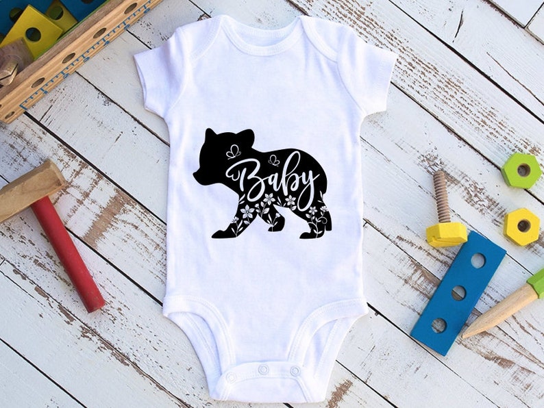 May include: White baby onesie with a black bear silhouette design. The bear is filled with floral patterns and the word "Baby" in a cursive font. The onesie is made of soft material and has snap closures.