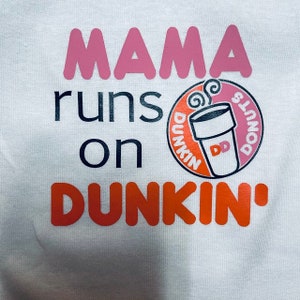 K&ouml;nnte beinhalten: Wei&szlig;es T-Shirt mit dem Text "Mama runs on Dunkin'" in Rosa, Schwarz und Orange. Ein Dunkin' Donuts Logo mit einem Kaffeebecher und Donuts befindet sich in der Mitte des Shirts.