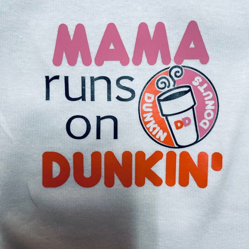 K&ouml;nnte beinhalten: Wei&szlig;es T-Shirt mit dem Text "Mama runs on Dunkin'" in Rosa, Schwarz und Orange. Ein Dunkin' Donuts Logo mit einem Kaffeebecher und Donuts befindet sich in der Mitte des Shirts.