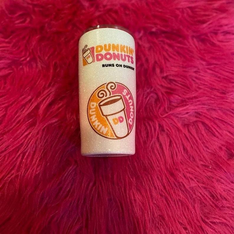 K&ouml;nnte beinhalten: Wei&szlig;er Glitzerbecher mit einem rosa und orangenen Dunkin' Donuts Logo. Das Logo lautet "Dunkin' Donuts" und "Runs on Dunkin'".