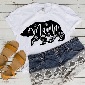 May include: White t-shirt with a black bear silhouette design that says "Mama" in cursive. The bear is decorated with floral and butterfly accents. The shirt is paired with denim shorts and brown sandals.