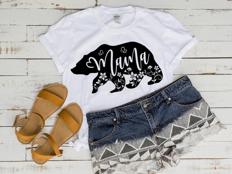 May include: White t-shirt with a black bear silhouette design that says "Mama" in cursive. The bear is decorated with floral and butterfly accents. The shirt is paired with denim shorts and brown sandals.