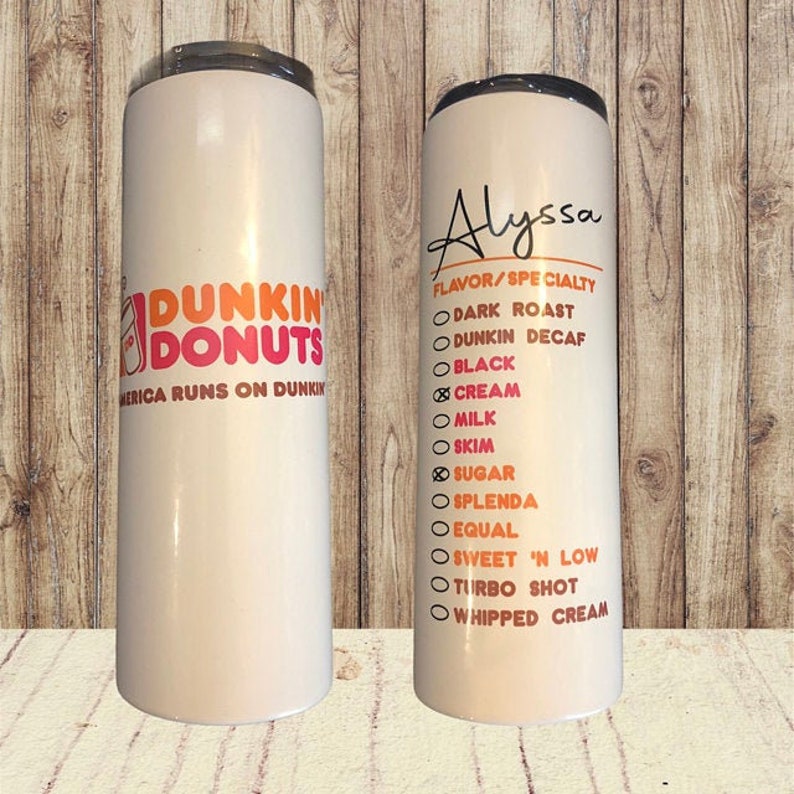 K&ouml;nnte beinhalten: Zwei wei&szlig;e Edelstahlbecher mit einem Dunkin' Donuts-Logo auf dem einen und einer Liste von Kaffeegeschmacksrichtungen und -optionen auf dem anderen. Der Becher rechts tr&auml;gt oben den Namen Alyssa.