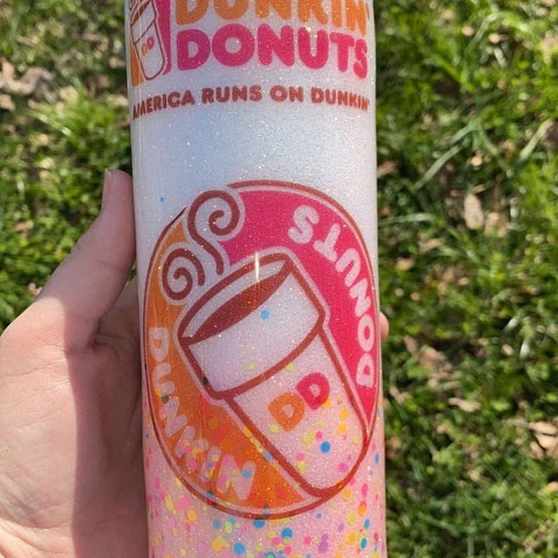 K&ouml;nnte beinhalten: Ein wei&szlig;er Becher mit einem rosafarbenen, orangen und wei&szlig;en Dunkin' Donuts Logo. Das Logo zeigt eine Tasse Kaffee und den Text "Dunkin' Donuts". Der Becher ist mit Glitzer bedeckt und hat einen wei&szlig;en Hintergrund.