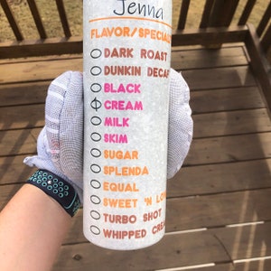 K&ouml;nnte beinhalten: Ein wei&szlig;er Becher mit einem schwarzen und orangen Design. Der Becher hat eine Liste von Kaffeeoptionen, darunter "Dark Roast", "Dunkin Decaf", "Black", "Cream", "Milk", "Skim", "Sugar", "Splenda", "Equal", "Sweet 'N Low", "Turbo Shot" und "Whipped Cream".