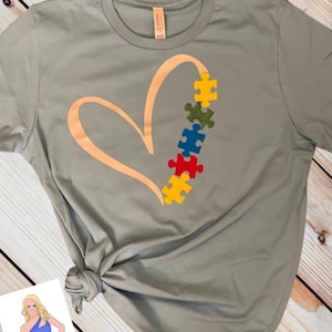 Puede incluir: Una camiseta gris con un dise&ntilde;o de coraz&oacute;n dorado y una fila de piezas de rompecabezas de colores. Las piezas del rompecabezas son rojas, amarillas, azules, verdes y amarillas.