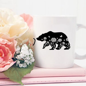 May include: White ceramic mug featuring a black silhouette of a bear filled with floral and butterfly designs. The mug is set on a stack of pink notebooks, with a bouquet of pink and white flowers in the background.