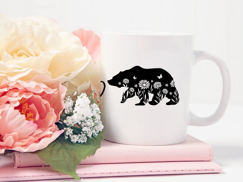 May include: White ceramic mug featuring a black silhouette of a bear filled with floral and butterfly designs. The mug is set on a stack of pink notebooks, with a bouquet of pink and white flowers in the background.