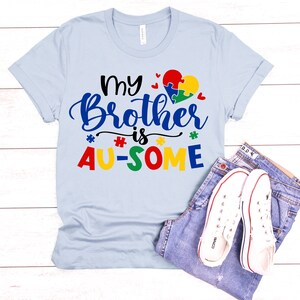 K&ouml;nnte beinhalten: Hellblaues T-Shirt mit dem Text "My Brother is AU-SOME" in bunten Buchstaben. Ein Puzzle-Herz befindet sich &uuml;ber dem Text.