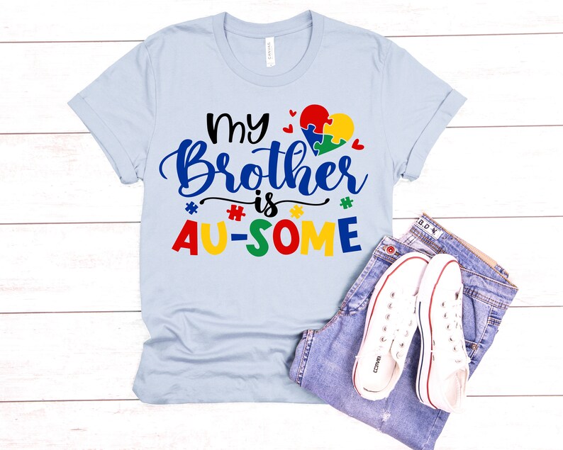 K&ouml;nnte beinhalten: Hellblaues T-Shirt mit dem Text "My Brother is AU-SOME" in bunten Buchstaben. Ein Puzzle-Herz befindet sich &uuml;ber dem Text.