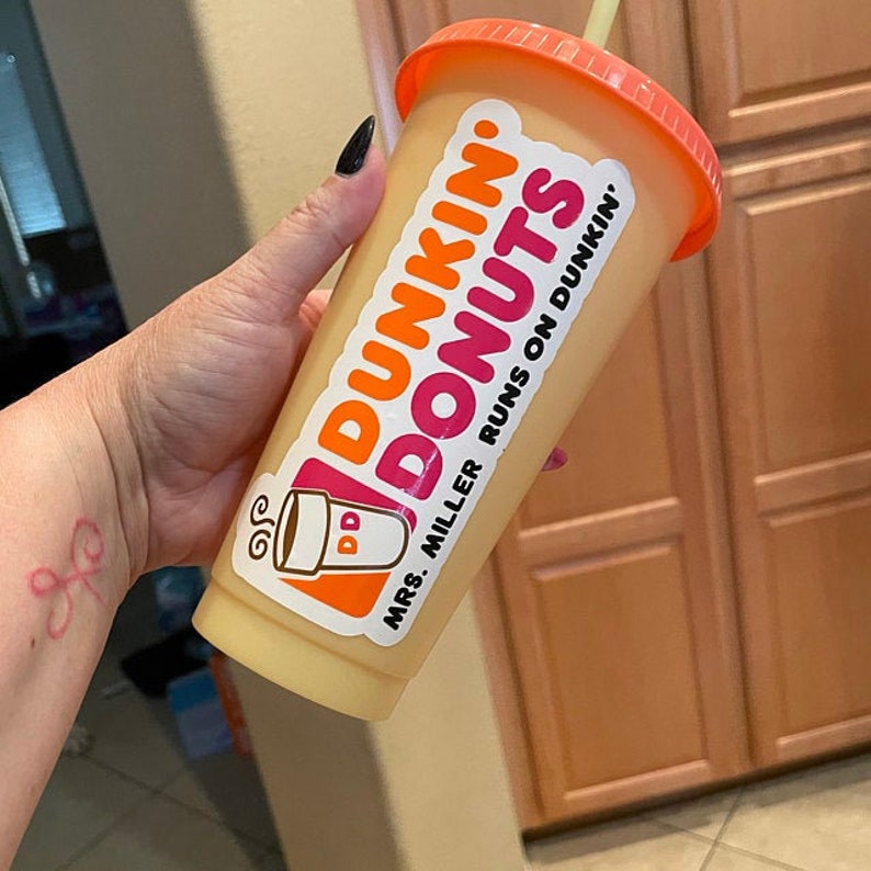K&ouml;nnte beinhalten: Ein durchsichtiger Plastikbecher mit einem rosafarbenen Deckel und einem Aufkleber mit der Aufschrift "Dunkin' Donuts" und "Mrs. Miller runs on Dunkin'" gef&uuml;llt mit einem hellgelben Getr&auml;nk.