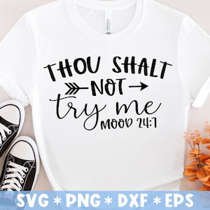 Op de afbeelding: Wit T-shirt met de zwarte tekst "THOU SHALT NOT try me MOOD 24:7". De afbeelding bevat zwart-witte sneakers. Een takje met oranje bloemen staat op de achtergrond.