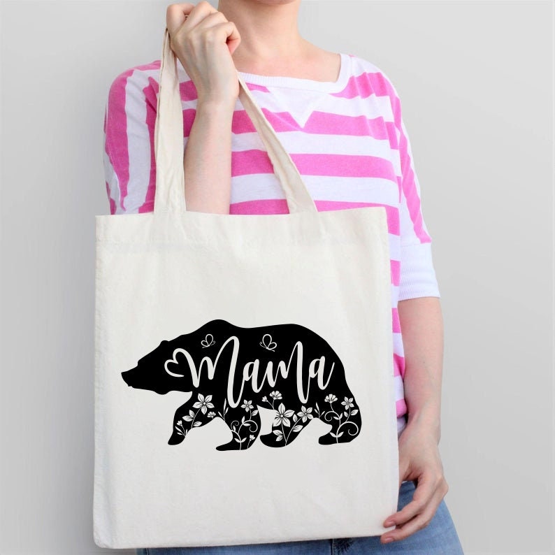 May include: Cream-colored tote bag featuring a black bear silhouette with the word "Mama" in cursive script. The bear design includes floral and butterfly accents, creating a nature-inspired theme. The bag is held by a person wearing a pink and white striped shirt.