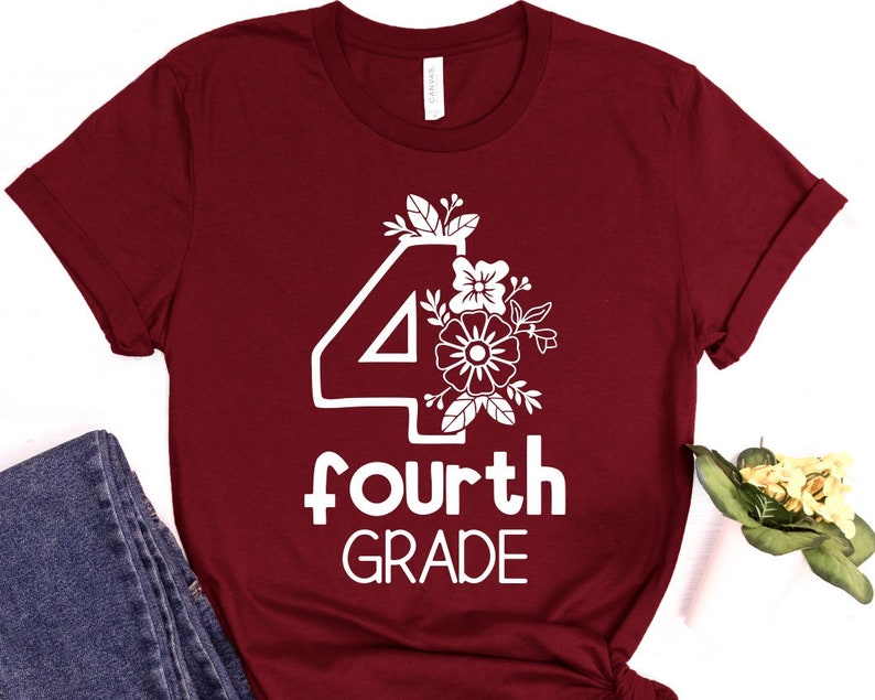 K&ouml;nnte beinhalten: Burgunderfarbenes T-Shirt mit wei&szlig;er Grafik. Die Grafik zeigt die Zahl 4, florale Akzente und die Worte "fourth GRADE" in fetter Schrift. Das T-Shirt ist gefaltet, eine Jeans ist in der Ecke sichtbar.