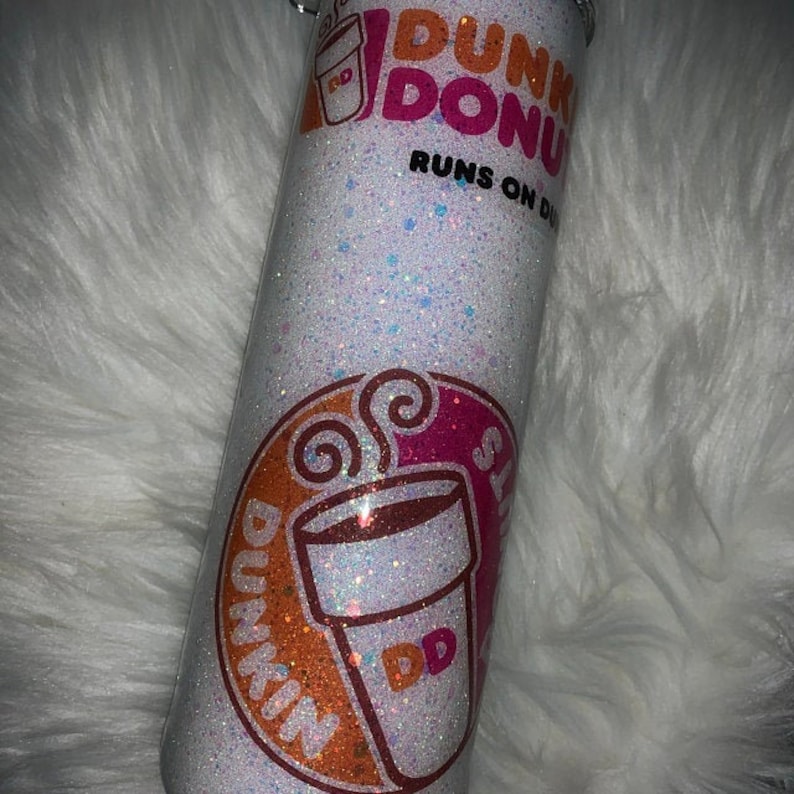 K&ouml;nnte beinhalten: Ein wei&szlig;er Glitzerbecher mit einem rosa und orangenen Dunkin' Donuts Logo und dem Text "Runs on Dunkin'" oben. Das Logo zeigt eine Kaffeetasse mit Dampf und einen Donut.