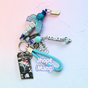 May include: A keychain featuring a braided light blue strap, a rectangular charm with a group photo and a cartoon character, and various beads. The beads spell out BTS and ARIRANG, accompanied by blue wing charms, a number 7 bead, and metal accents.