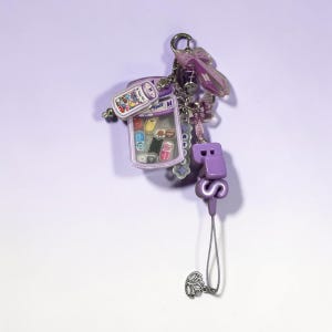 May include: A purple-themed keychain featuring a miniature jar charm filled with colorful character icons, a purple bow charm, and large purple block letters spelling BTS. The accessory includes a silver metal ring and a small lotus-shaped charm.