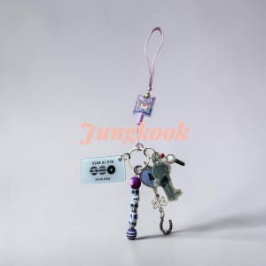 May include: A purple lanyard keychain featuring a rectangular charm with the text BTS IS BACK, a purple bead strand spelling BTS 7, a heart-shaped charm, a silhouette figure, a decorative knot, and a small horseshoe charm on a metal ring.