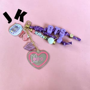 May include: A purple cord keychain featuring a heart-shaped charm with a pink border and the BTS logo. It includes letter beads spelling BTS and ARIRANG, a small digital pet-style charm with a photo, and a purple acrylic charm labeled Jungkook.