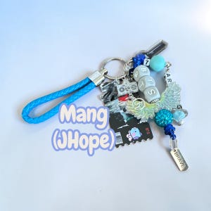May include: A keychain featuring a blue braided wrist strap attached to a metal ring. It includes various charms: a clear wing, a blue glitter bead, a silver tag reading BRAVE, and white cube beads spelling BTS. A black rectangular charm displays a cartoon character.