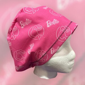 May include: Pink surgical scrub hat with a white Barbie logo pattern. The hat has a black elastic band.