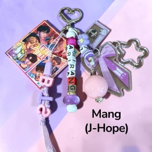 May include: A multi-strand keychain featuring a square charm with a BTS group photo, a purple ribbon charm with a BTS logo, and a star-shaped clasp. It includes letter beads spelling ARIRANG, pink and purple spherical beads, and a BTS letter charm.