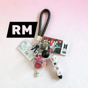 May include: A keychain featuring a black braided strap, a rectangular charm with a group photo of seven individuals, a white ticket-shaped charm with the BTS logo, and a clear charm with text. Includes beads spelling ARMY and a pink heart charm.