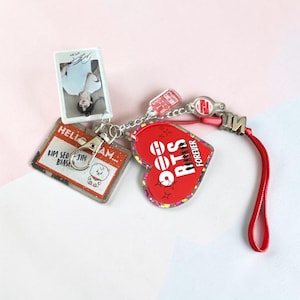 May include: A multi-charm keychain featuring a rectangular name tag with the text HELLO I AM KIM SEOK-JIN BIASED, a small cartoon character, a vertical photo card of a person, a red heart-shaped charm with the text BTS FOREVER, and a red strap.