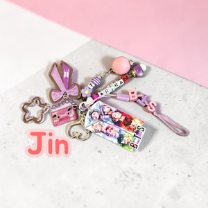 May include: A decorative keychain featuring a rectangular charm with a collage of seven individuals, a purple ribbon charm with the BTS logo, a pink cassette tape charm, and a string of beads spelling ARIRANG and BTS with a large pink bead.