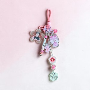 May include: A decorative keychain featuring a pink cord, a flower-shaped charm with a portrait, a pink bead strand labeled ARIRANG, a purple puzzle piece charm with I-HOPE text, a pink bear character charm, and various floral beads.