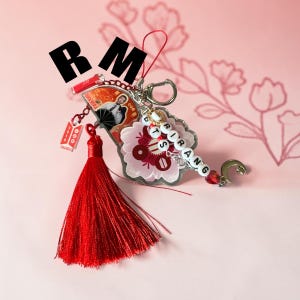 May include: A decorative keychain featuring a large red tassel, a fan-shaped charm with a portrait, a flower-shaped acrylic piece, and white letter beads spelling ARIRANG. It includes a silver metal clasp, a small crescent moon, and red accents.