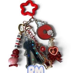 May include: A red star-shaped keychain featuring multiple charms, including a clear acrylic figure of a person, a white bead strand spelling BTS, a red circular charm with white cartoon characters, a red heart, a red bow, and a small red lightstick.