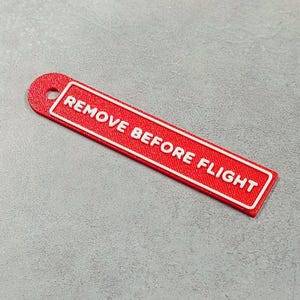 Puede incluir: Una etiqueta rectangular roja con el texto "REMOVE BEFORE FLIGHT" en blanco. La etiqueta tiene un borde blanco y un agujero en la parte superior para sujetarla. La etiqueta está sobre una superficie gris.