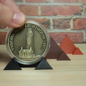 May include: A gold-colored coin featuring the Yaquina Head Lighthouse, dated 1873, displayed in a clear case. The coin rests on a black triangular stand, with brown and red triangular shapes in the background.