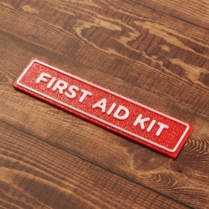 May include: A rectangular red sign with the words "FIRST AID KIT" in white, raised lettering. The sign has a white border and is set against a wooden background.
