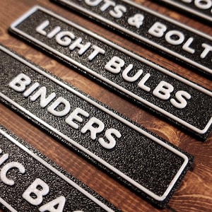 Könnte beinhalten: Nahaufnahme von mehreren schwarz-weißen Etiketten mit erhabenen weißen Buchstaben. Die Etiketten lauten "LIGHT BULBS" und "BINDERS". Die Etiketten haben eine strukturierte Oberfläche und einen weißen Rand. Die Etiketten dienen wahrscheinlich der Organisation.