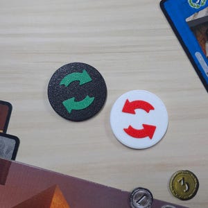 May include: Two circular game tokens. One is black with green arrows, the other is white with red arrows. The arrows are in a circular motion. The tokens are on a light brown surface with other game pieces.