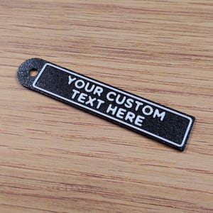Puede incluir: Un llavero rectangular negro con el texto "YOUR CUSTOM TEXT HERE" en blanco. El llavero tiene un borde blanco y un pequeño agujero en la parte superior para sujetarlo. El fondo es una superficie de madera marrón claro.