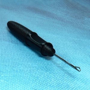 May include: A black crochet hook with a textured handle and a metal hook end. The hook is designed for pulling yarn through fabric loops to create various crochet projects. The tool is set against a blue background.