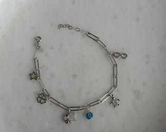 May include: A silver-toned chain link bracelet featuring multiple charms including a star, a four-leaf clover, a textured animal figure, a small blue and white evil eye bead, a sea turtle, and an infinity symbol, finished with a lobster clasp.